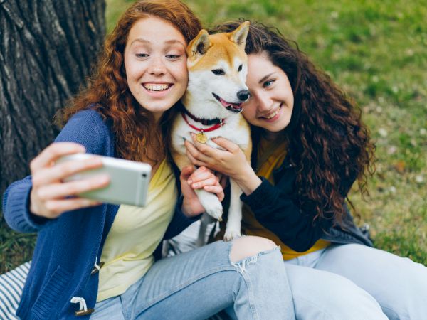 Two people are sitting on the grass, taking a selfie with a dog between them. They are smiling and look happy together.
