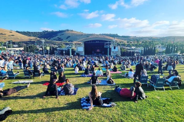 People sitting on a grassy field, some on blankets, others in chairs, facing an outdoor stage with hills in the background under a bright blue sky.