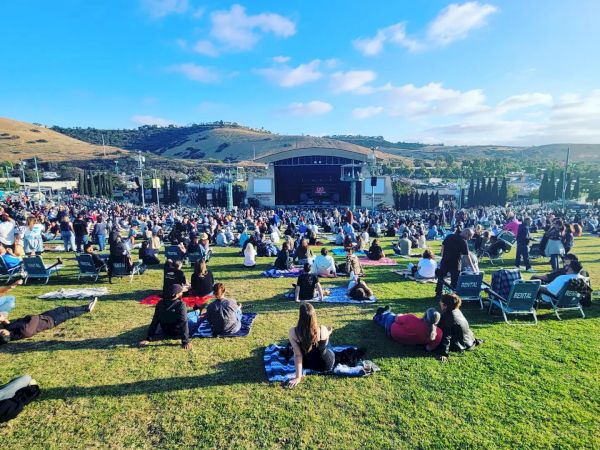 People sitting on a grassy field, some on blankets, others in chairs, facing an outdoor stage with hills in the background under a bright blue sky.
