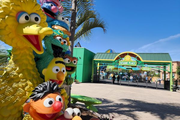 A colorful sculpture of Sesame Street characters stands near the entrance of Sesame Place, with palm trees and a blue sky in the background.