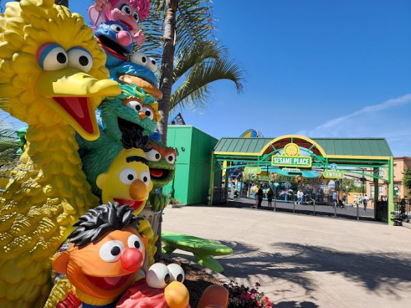 A colorful sculpture of Sesame Street characters stands near the entrance of Sesame Place, with palm trees and a blue sky in the background.