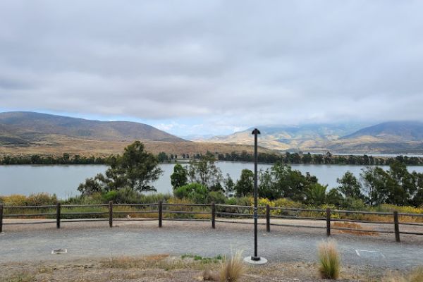 A scenic view of a lake with mountains and cloudy skies in the background, bordered by trees and a wooden fence with a lamp post in the foreground.