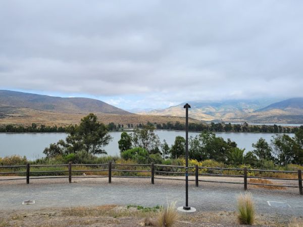 A scenic view of a lake with mountains and cloudy skies in the background, bordered by trees and a wooden fence with a lamp post in the foreground.