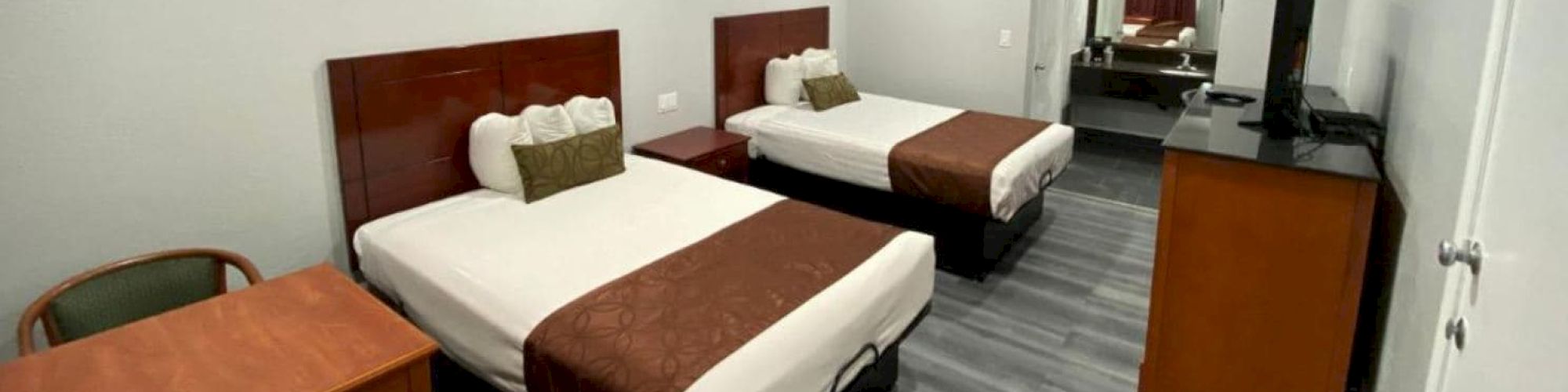 The image shows a hotel room with two beds, a small table with chairs, a TV, and a dresser on a wood floor. It has a simple, modern look.