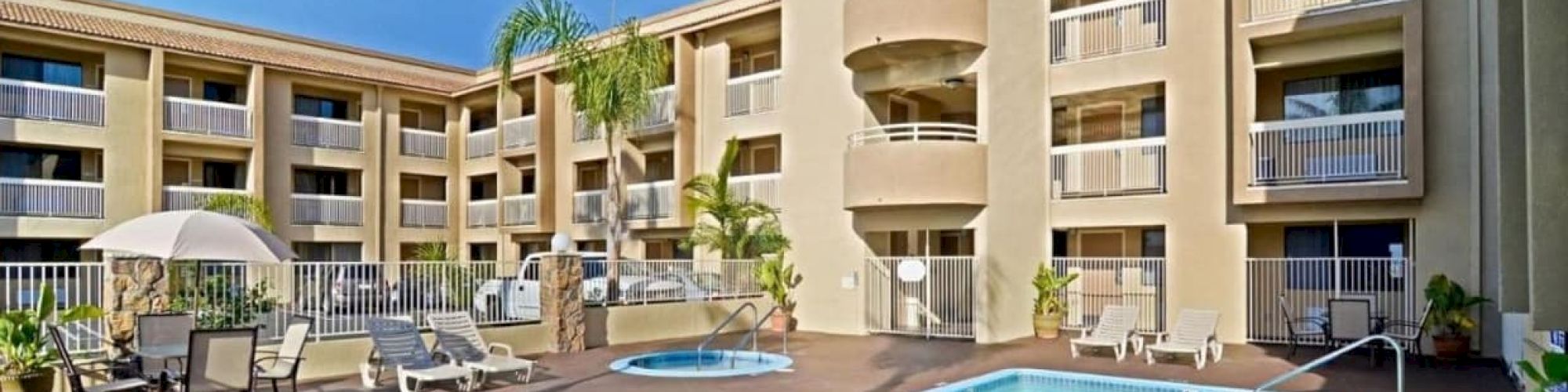 An outdoor pool area with lounge chairs and tables, surrounded by a three-story building with balconies and palm trees, on a sunny day.