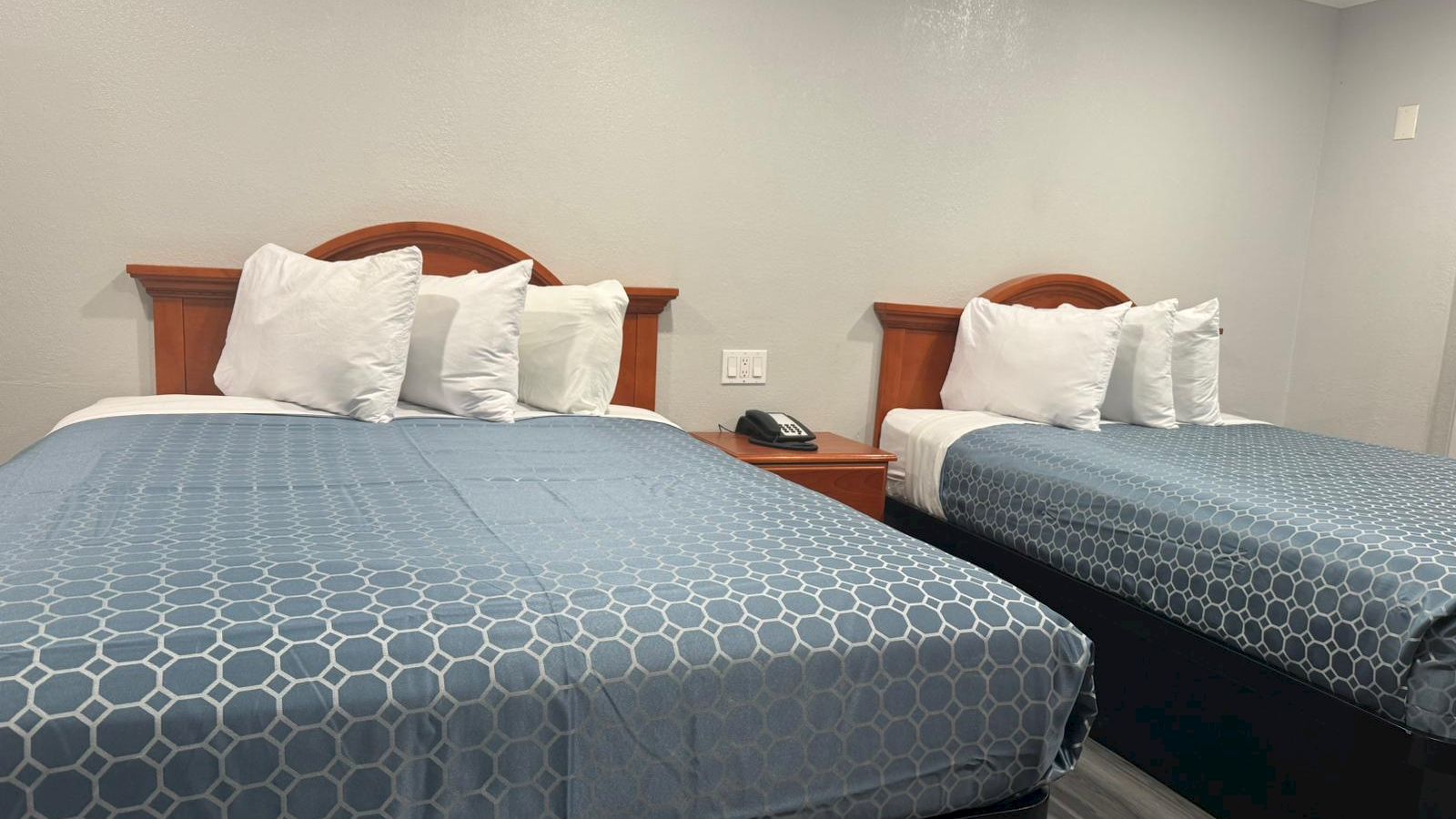 The image shows a hotel room with two beds, blue bedspreads, white pillows, a nightstand, a phone, and white walls.