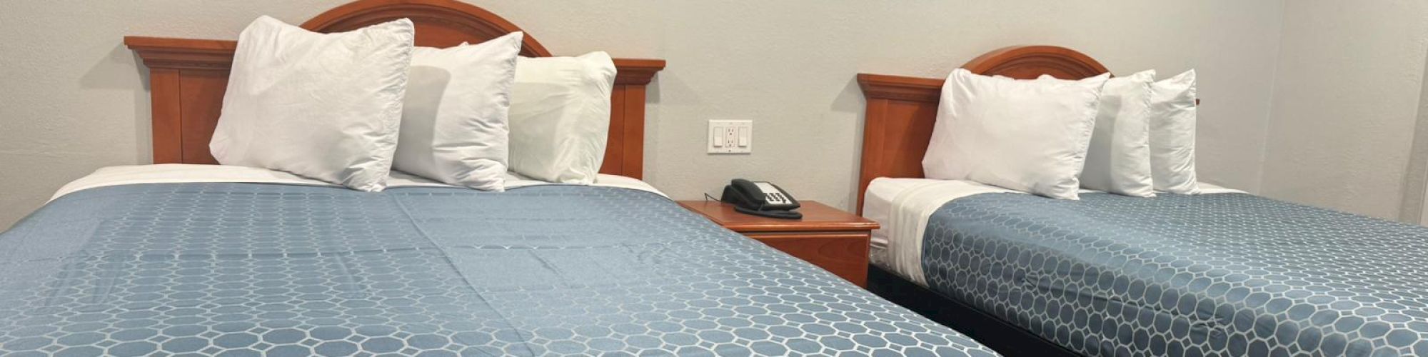 The image shows a hotel room with two beds, blue bedspreads, white pillows, a nightstand, a phone, and white walls.