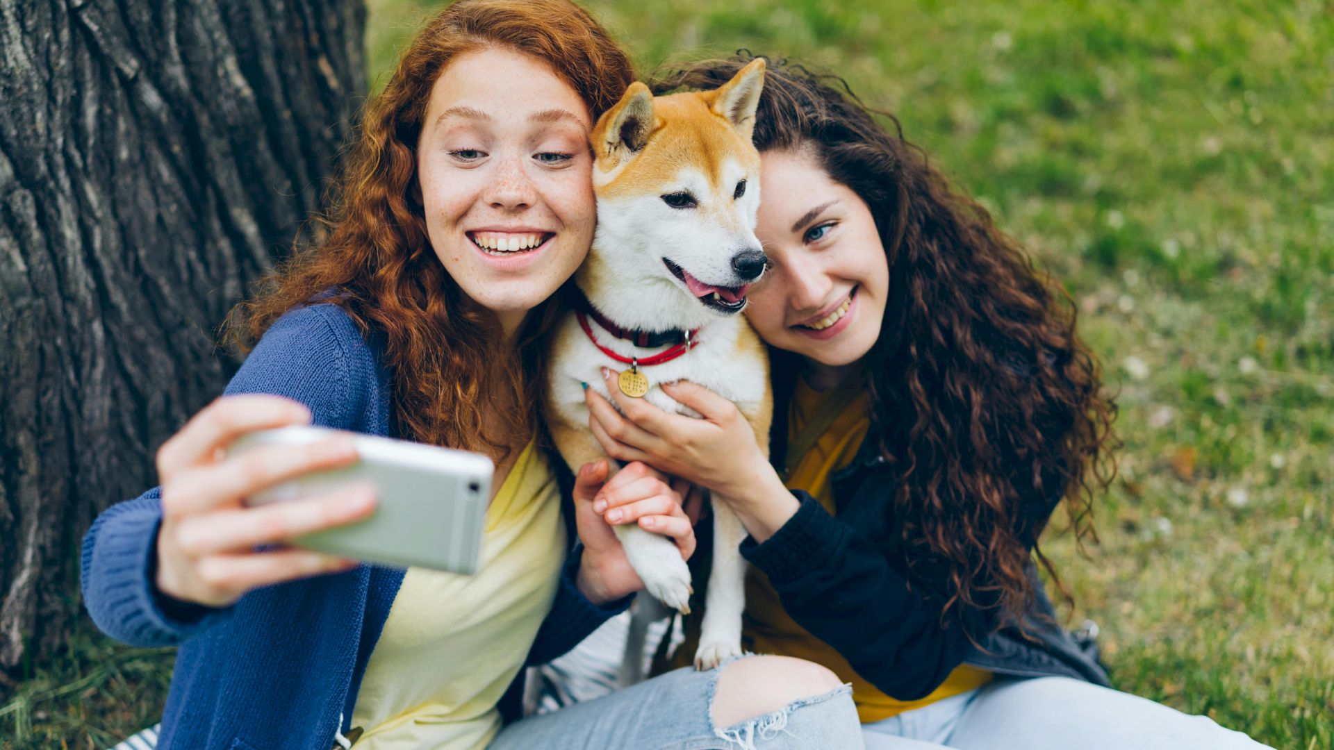 Two women sitting on the ground, taking a selfie with a dog, all smiling and looking happy.