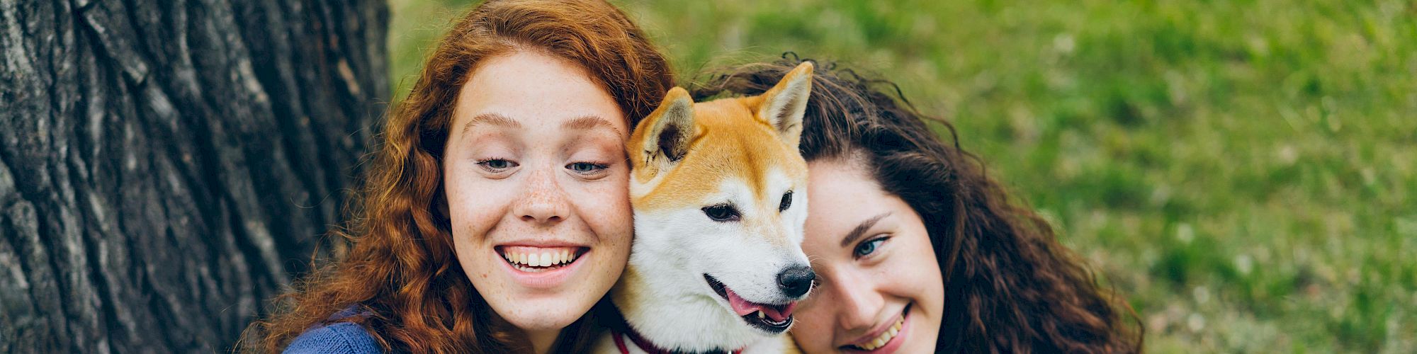 Two women sitting on the ground, taking a selfie with a dog, all smiling and looking happy.