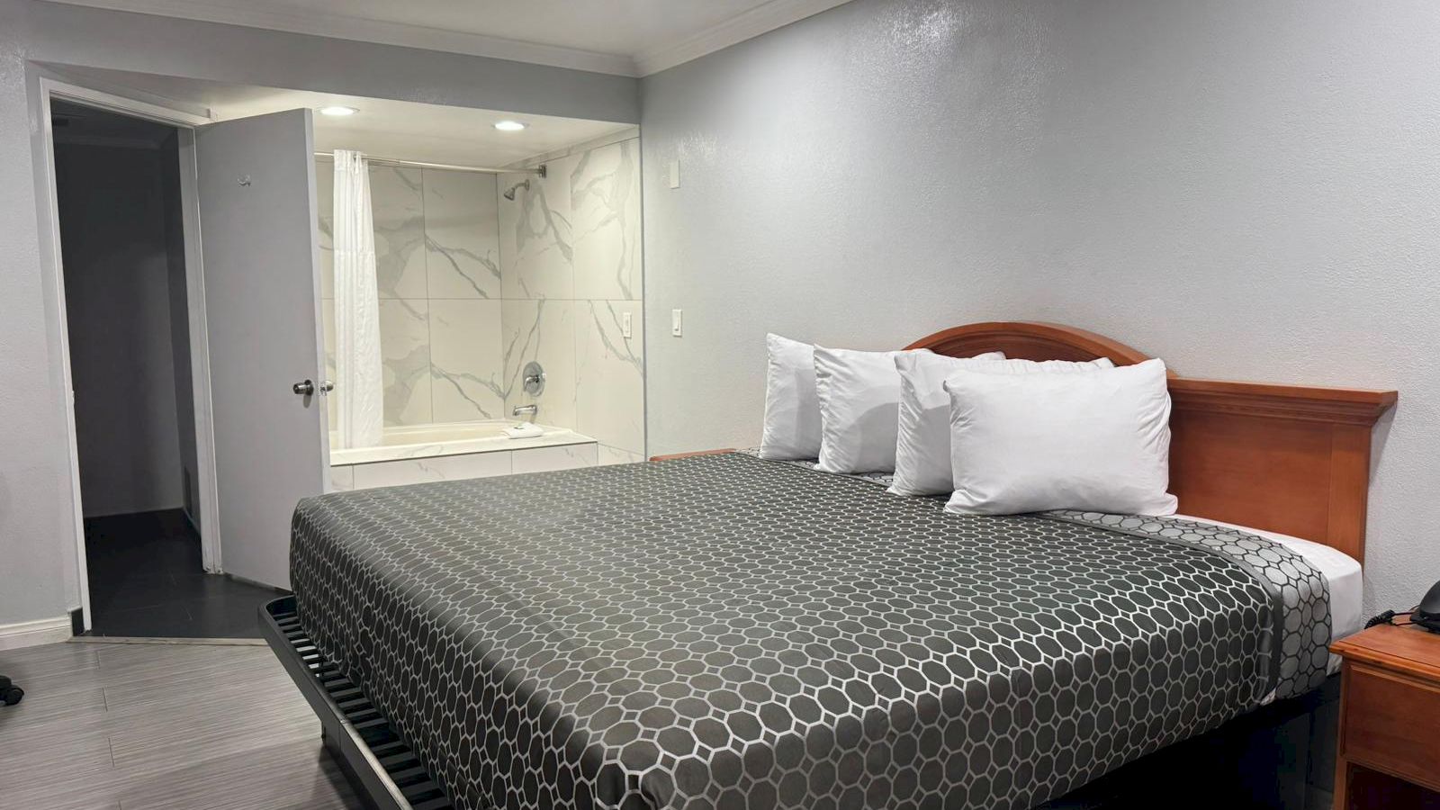 A hotel room with a king-size bed, four pillows, a nightstand, and an open bathroom area with a bathtub.