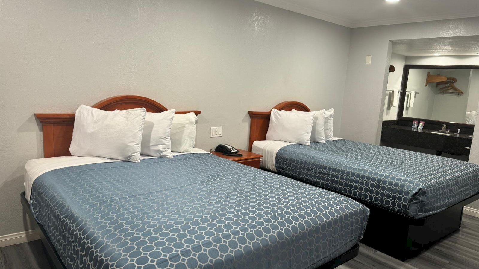 A hotel room with two double beds, white pillows, blue bedspreads, a nightstand, and a large mirror with a sink in the background.