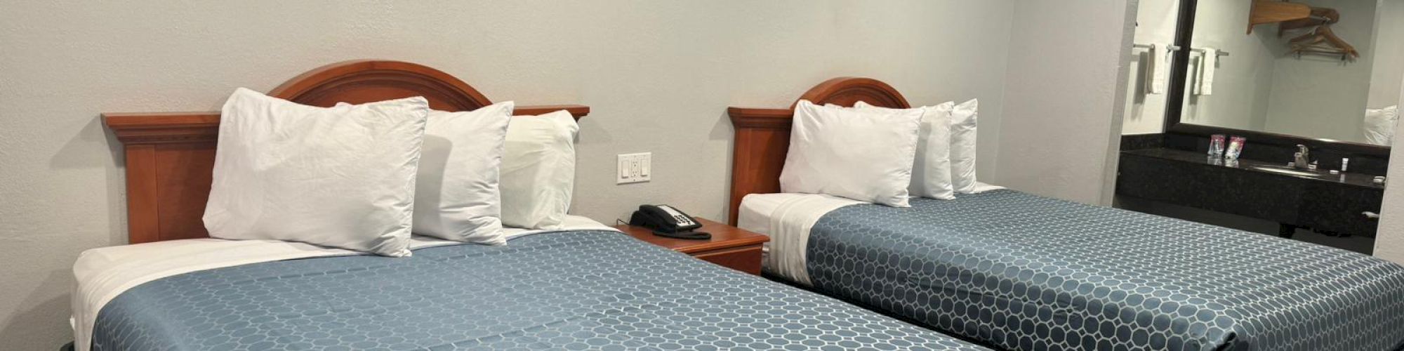 A hotel room with two double beds, white pillows, blue bedspreads, a nightstand, and a large mirror with a sink in the background.