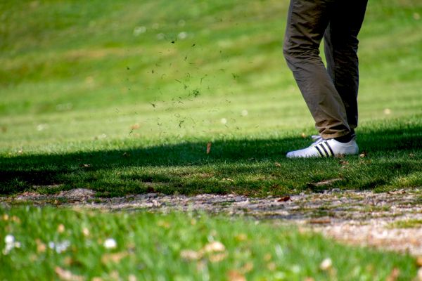 A person is seen from the waist down, wearing brown pants and white Adidas sneakers, on a grassy area likely on a golf course.