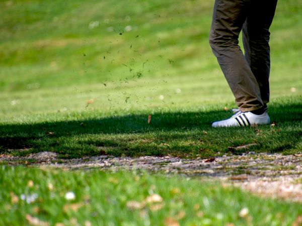 A person is seen from the waist down, wearing brown pants and white Adidas sneakers, on a grassy area likely on a golf course.