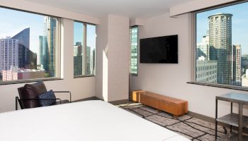 A modern room with a bed, chair, TV, and windows providing a cityscape view featuring tall buildings in the background.