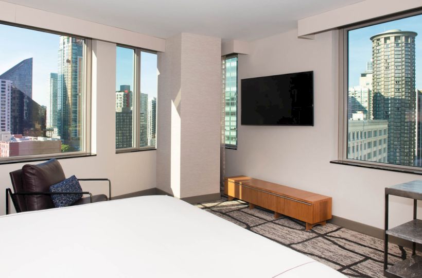 A modern room with a bed, chair, TV, and windows providing a cityscape view featuring tall buildings in the background.