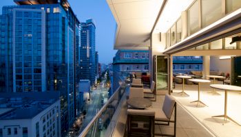 This image shows a modern cityscape with tall buildings and a terrace featuring outdoor seating, taken at twilight.