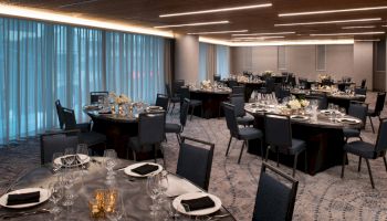 A banquet hall with round tables set with black napkins, glassware, and floral centerpieces is arranged under modern lighting fixtures.