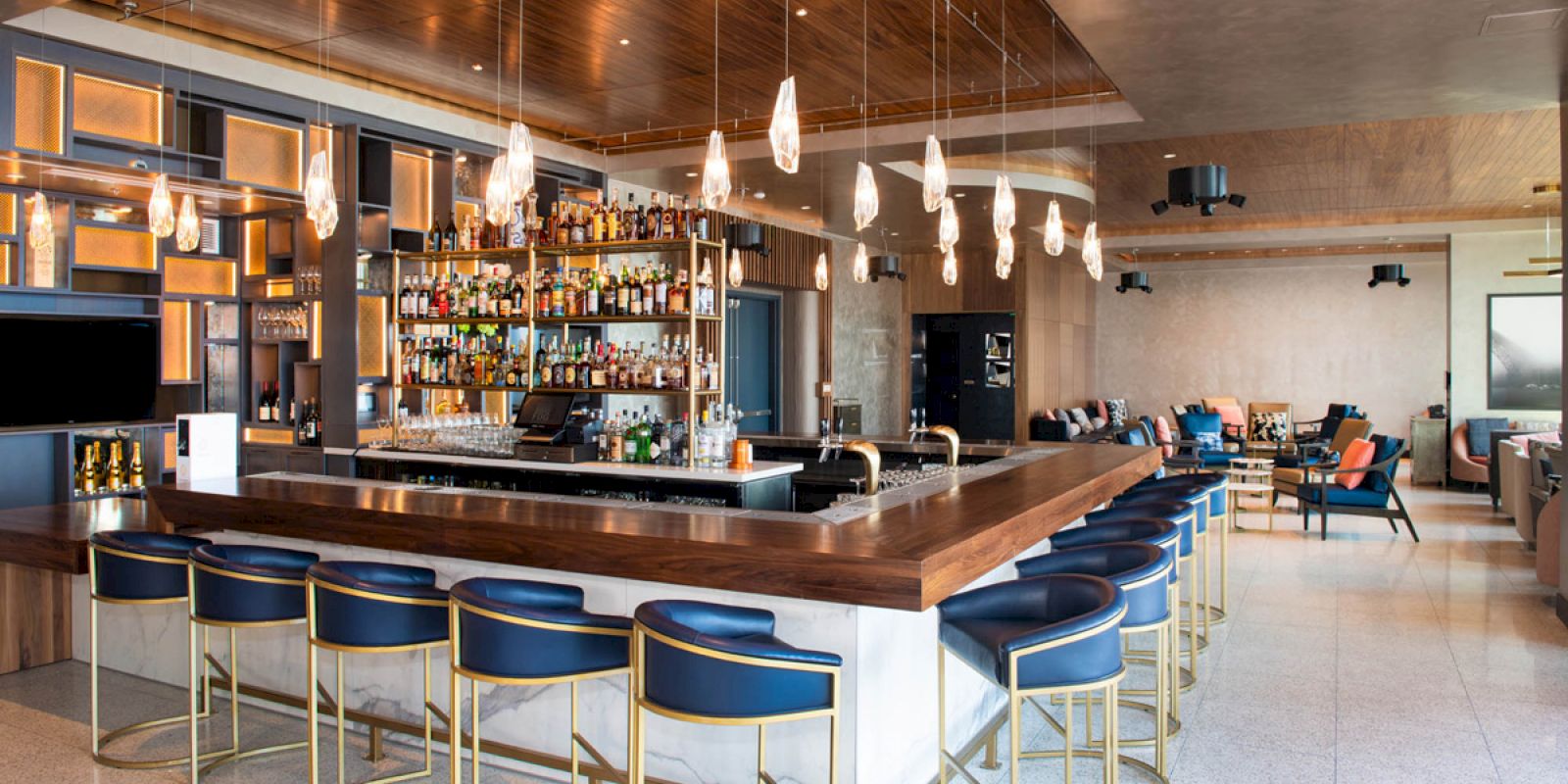 A modern bar with a wooden counter, blue and gold barstools, hanging lights, and a well-stocked liquor display, with seating areas in the background.
