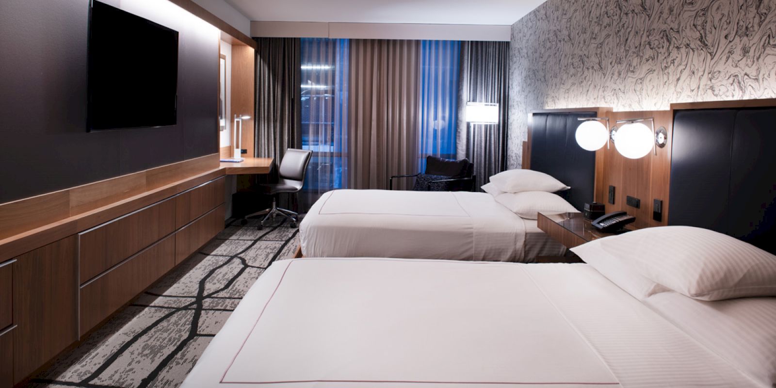 A modern hotel room with two neatly made beds, a desk, chair, wall-mounted TV, and a window with curtains. The room features a minimalistic design.