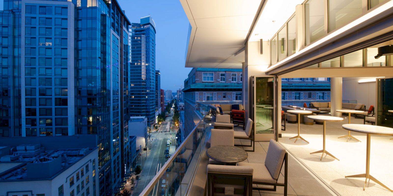 A modern balcony with outdoor seating overlooks a city street lined with tall buildings in the evening.