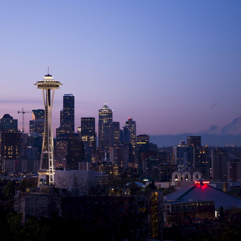 The image shows a cityscape at dusk, featuring the Space Needle prominently, with a mountainous backdrop and a mix of modern and older buildings.
