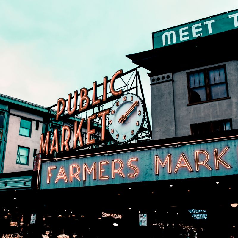 Image of a public farmers market with neon signs and a clock. "Public Market" and "Farmers Market" are prominently displayed on the buildings.