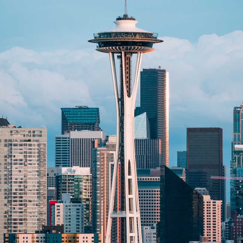 The image shows the Space Needle, a prominent observation tower in Seattle, with the city's skyline featuring modern skyscrapers in the background.
