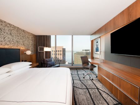 A modern hotel room with a large bed, a desk with a chair, a big TV, and a floor-to-ceiling window offering a city and water view.