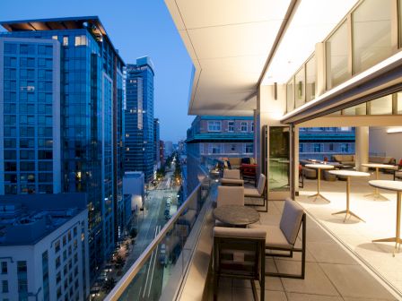 A modern urban terrace offers a view of the cityscape at dusk, featuring outdoor seating, tables, and adjacent tall buildings illuminated by city lights.