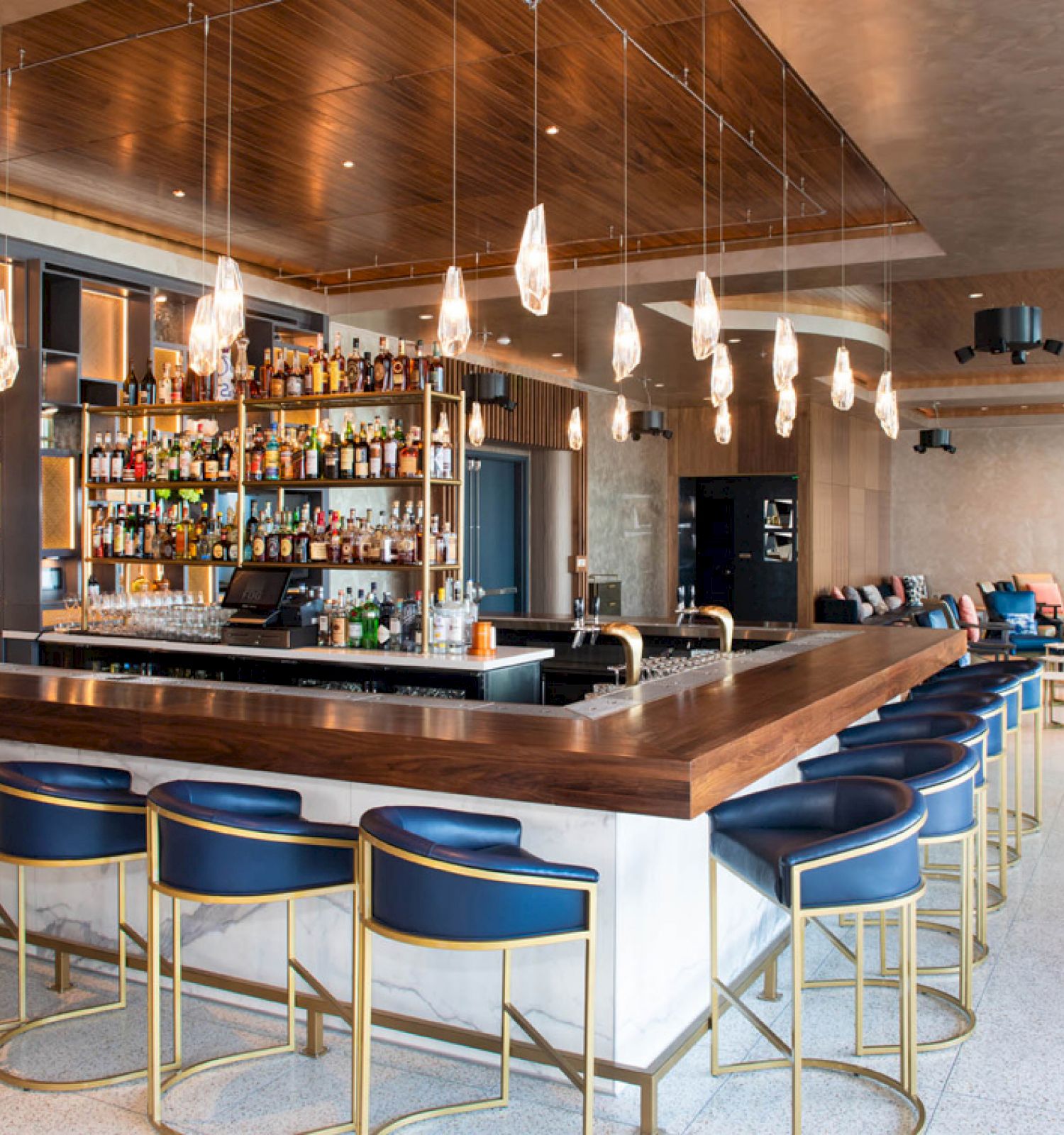 This image shows a modern bar with blue seating, a variety of drinks on shelves, pendant lights, and a lounge area in the background.