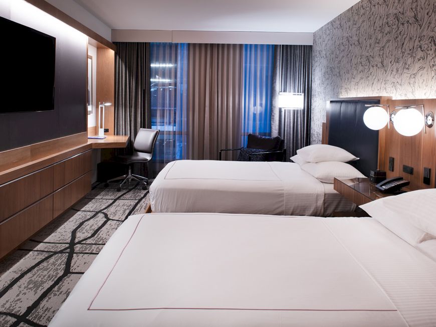 A modern hotel room with two beds, wall-mounted TV, desk with chair, bedside lamps, and large windows with curtains, creating a cozy atmosphere.