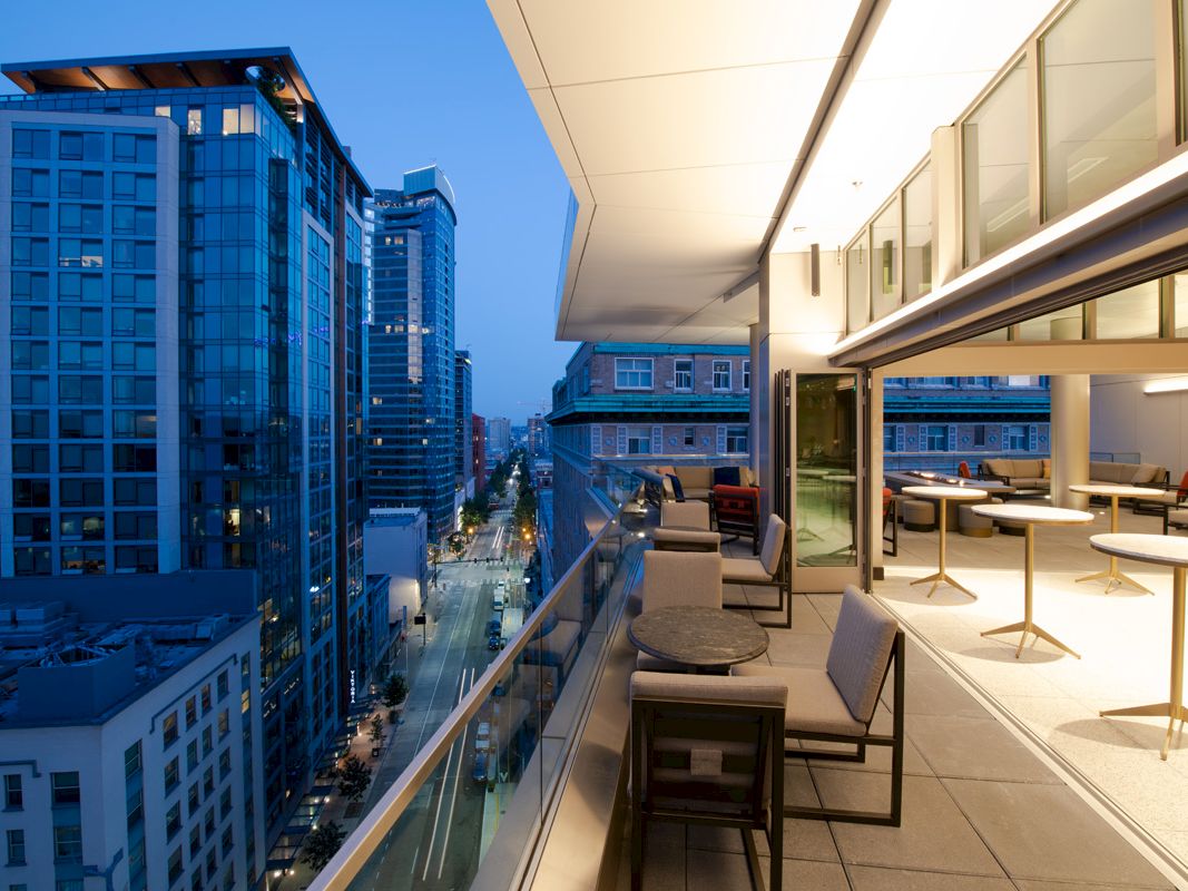Modern cityscape viewed from a high balcony at dusk, featuring high-rise buildings and a furnished outdoor seating area.