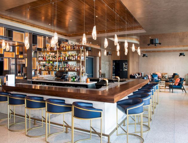 The image shows a modern bar with blue stools, a wooden counter, and a well-stocked liquor display. There are lounge areas in the background.