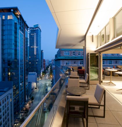A modern balcony at dusk overlooks a city street with tall buildings. There are tables, chairs, and bright interior lighting visible inside.
