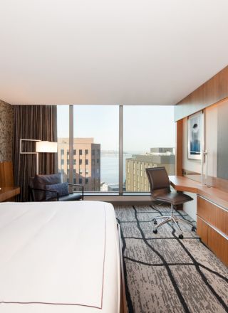 A modern hotel room with a large bed, desk, chair, television, and a window with a view of buildings and water.