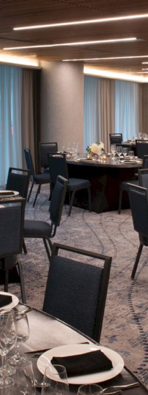 A banquet hall is set up with round tables covered in black tablecloths, each adorned with glassware, napkins, and floral centerpieces.