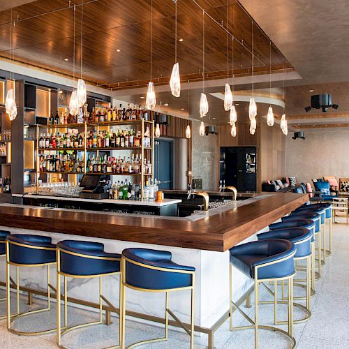 A modern bar with blue and gold stools, a wooden counter, a fully stocked back bar, pendant lights, and a seating area in the background.