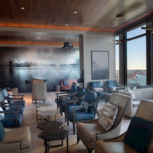 The image shows a modern lounge area with comfortable seating, large windows offering a waterfront view, and skyline artwork on the wall.