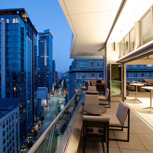 A modern cityscape at dusk featuring a balcony with seating and tables overlooking a busy urban street lined with buildings.