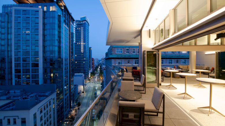 A modern city balcony at dusk with a view of tall buildings, street, and seating areas, illuminated by warm lights from inside.