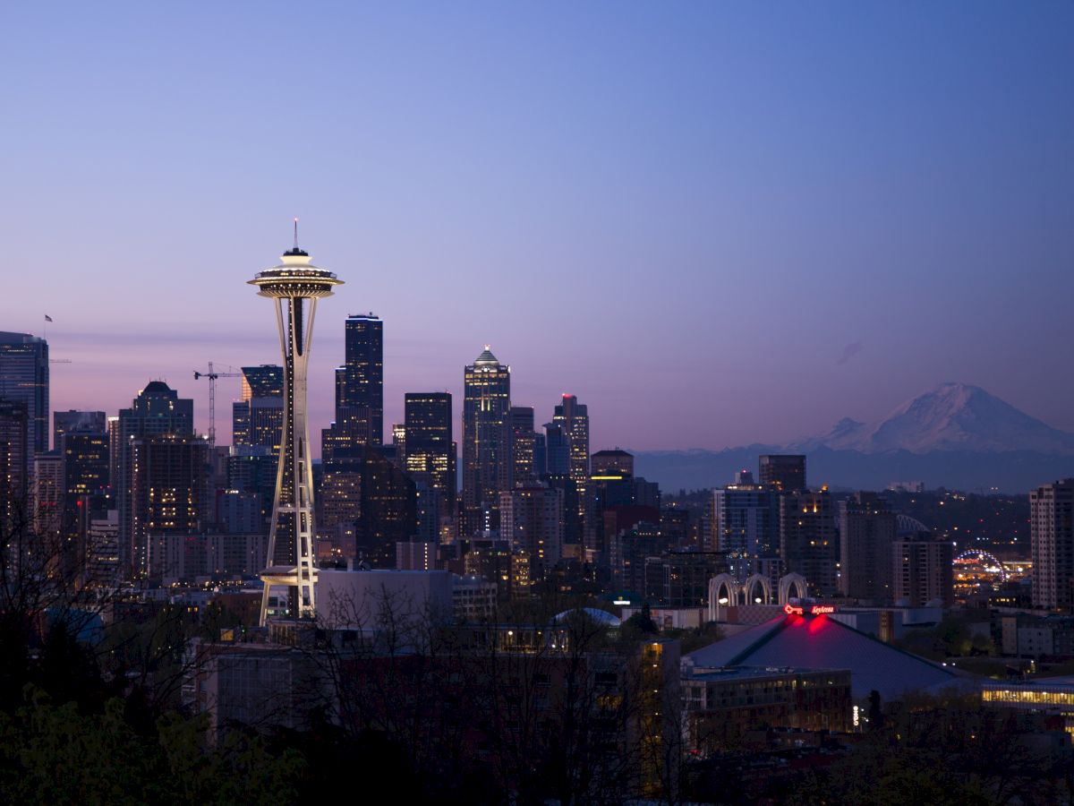 The image shows Seattle's skyline at dusk with the Space Needle prominently lit and Mount Rainier in the backdrop under a twilight sky.