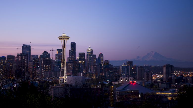 The image shows Seattle's skyline at dusk with the Space Needle prominently lit and Mount Rainier in the backdrop under a twilight sky.