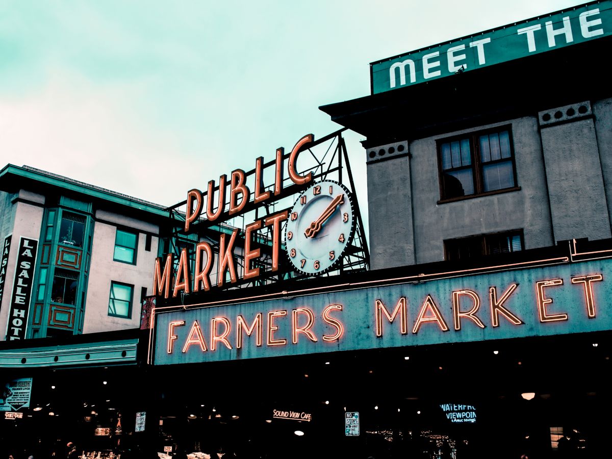 This image shows the exterior of a public farmers market with neon signs and buildings in the background.