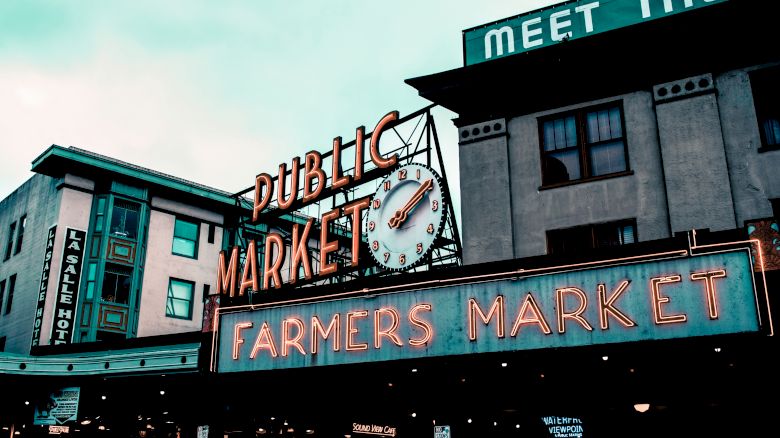 This image shows the exterior of a public farmers market with neon signs and buildings in the background.