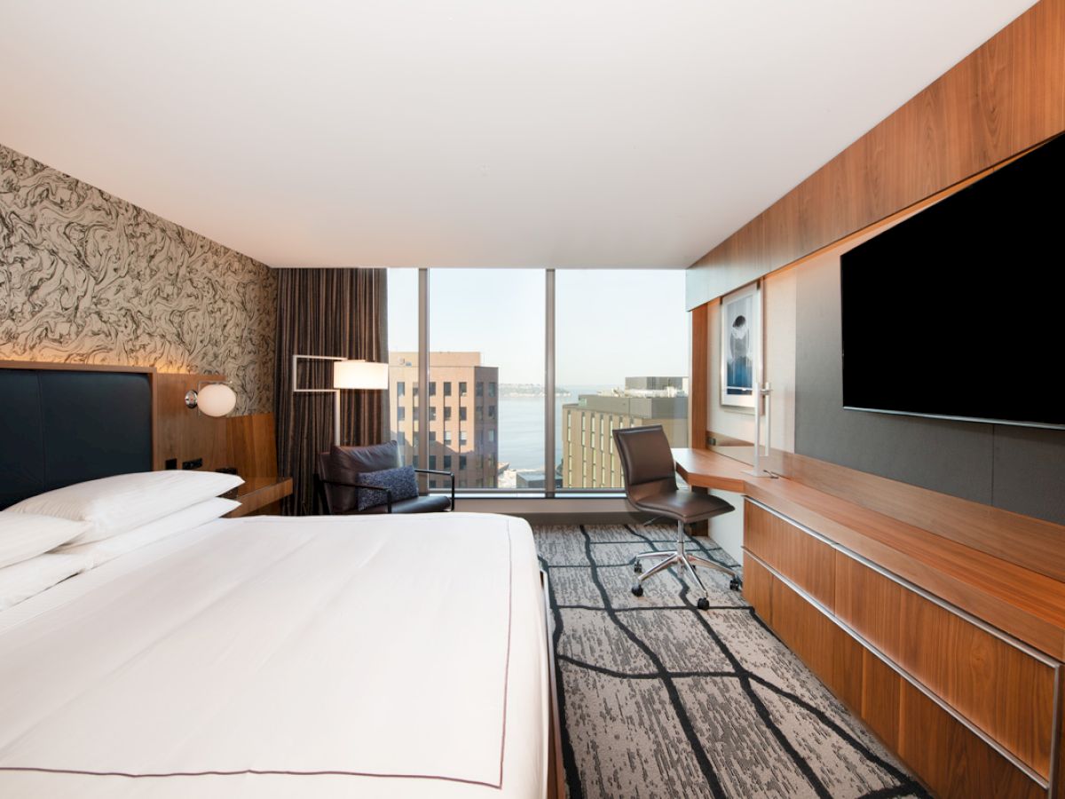 The image shows a modern hotel room with a large bed, a wall-mounted TV, a desk, and a chair by a window offering a cityscape view.
