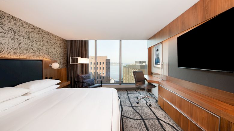 The image shows a modern hotel room with a large bed, a wall-mounted TV, a desk, and a chair by a window offering a cityscape view.
