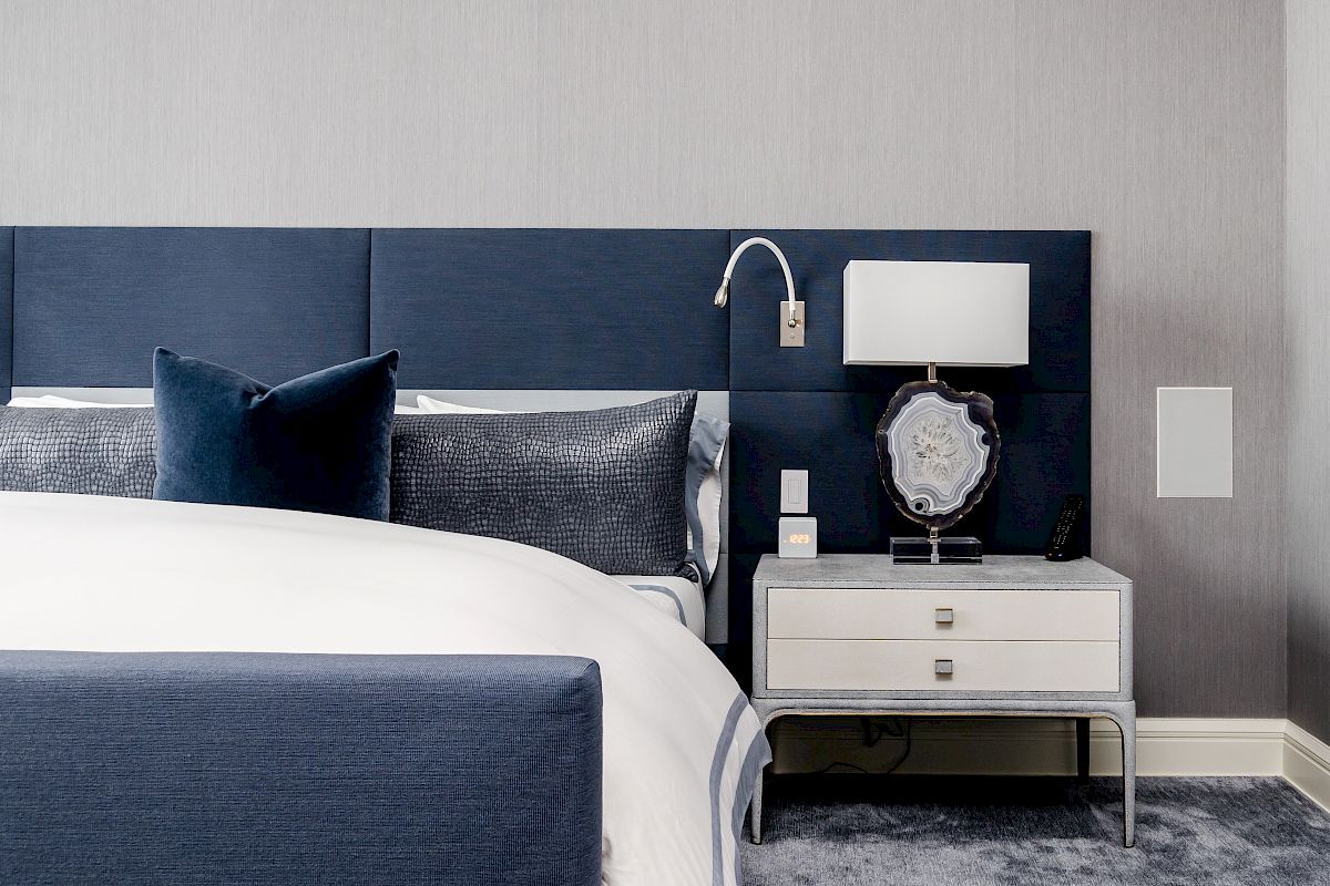 A modern bedroom with a blue upholstered headboard, white and blue bedding, a nightstand with a lamp, and a textured gray carpeted floor.