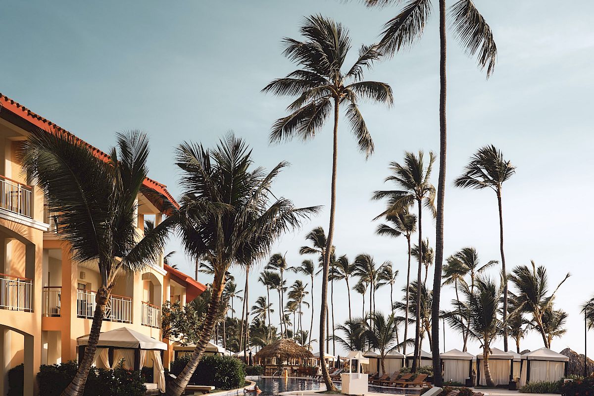 A serene poolside scene featuring lounge chairs, tall palm trees, and a building with balconies under a partly cloudy sky, exuding a tropical vibe.