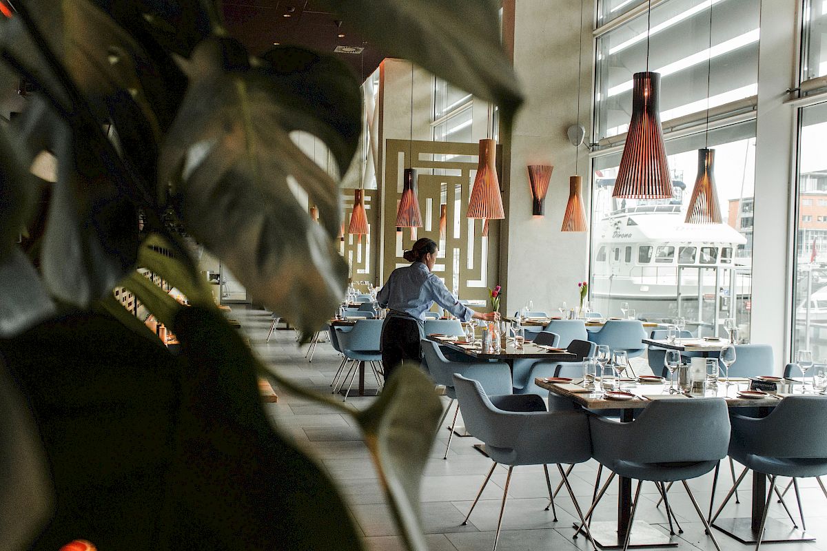 A modern restaurant with large windows, stylish lighting, and chairs neatly arranged; staff member is preparing the tables in the background.